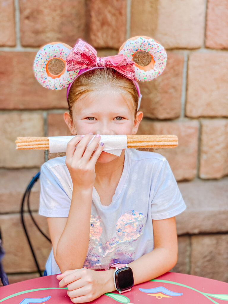a child wearing donut Minnie Mouse ears holding a foot long churro in front of her face like a mustache
